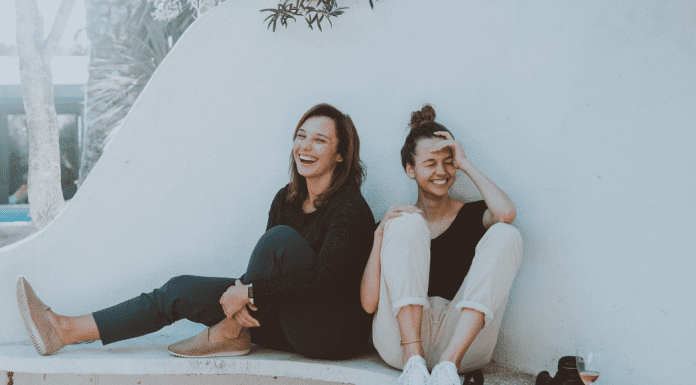 Two women laughing outside sitting against a white wall. Loving lately. Des Moines Mom
