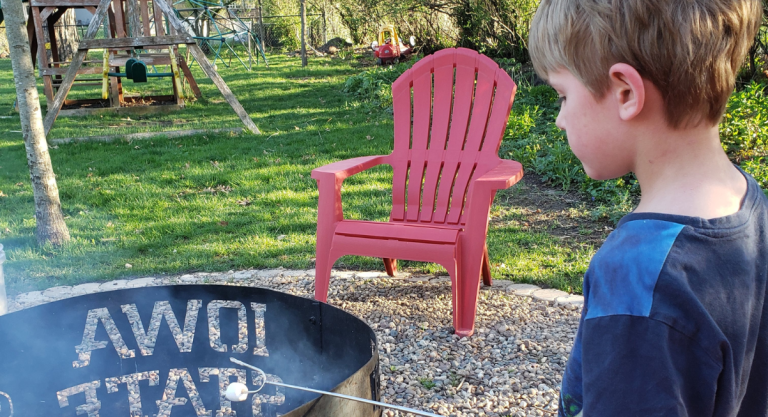 Boy with short hair and light skin roasting a marshmallow on a stick over a fire pit in a backyard with green grass.