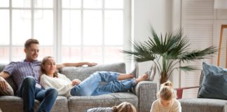 Children sister and brother playing drawing together on floor while young parents relaxing at home on sofa, little boy girl having fun, friendship between siblings, family leisure time in living room